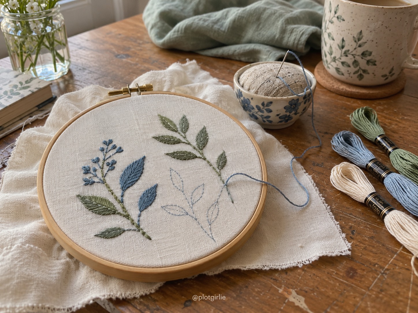 An unfinished botanical embroidery hoop with blue and sage thread on a wooden table